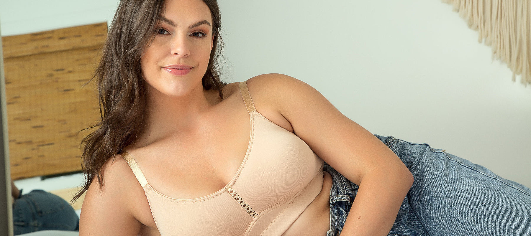 Woman wearing a beige bralette and blue jeans in a bedroom setting