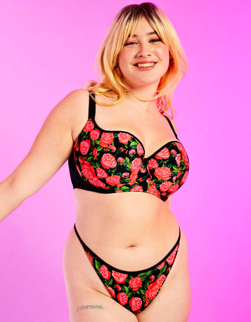 A woman modeling a two-piece lingerie set with a black background and a vibrant pink and green rose pattern, set against a solid pink studio background