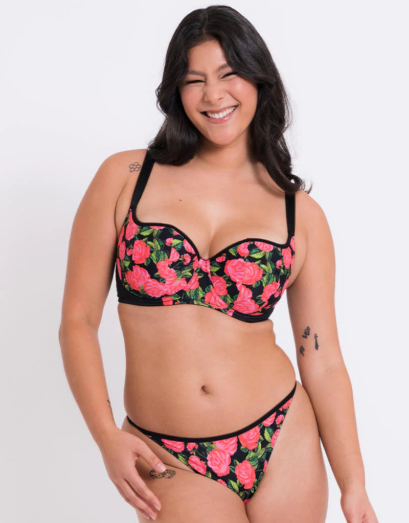 A woman modeling a two-piece bikini set with a black background and a vibrant pink rose floral print, set against a plain light gray studio background.