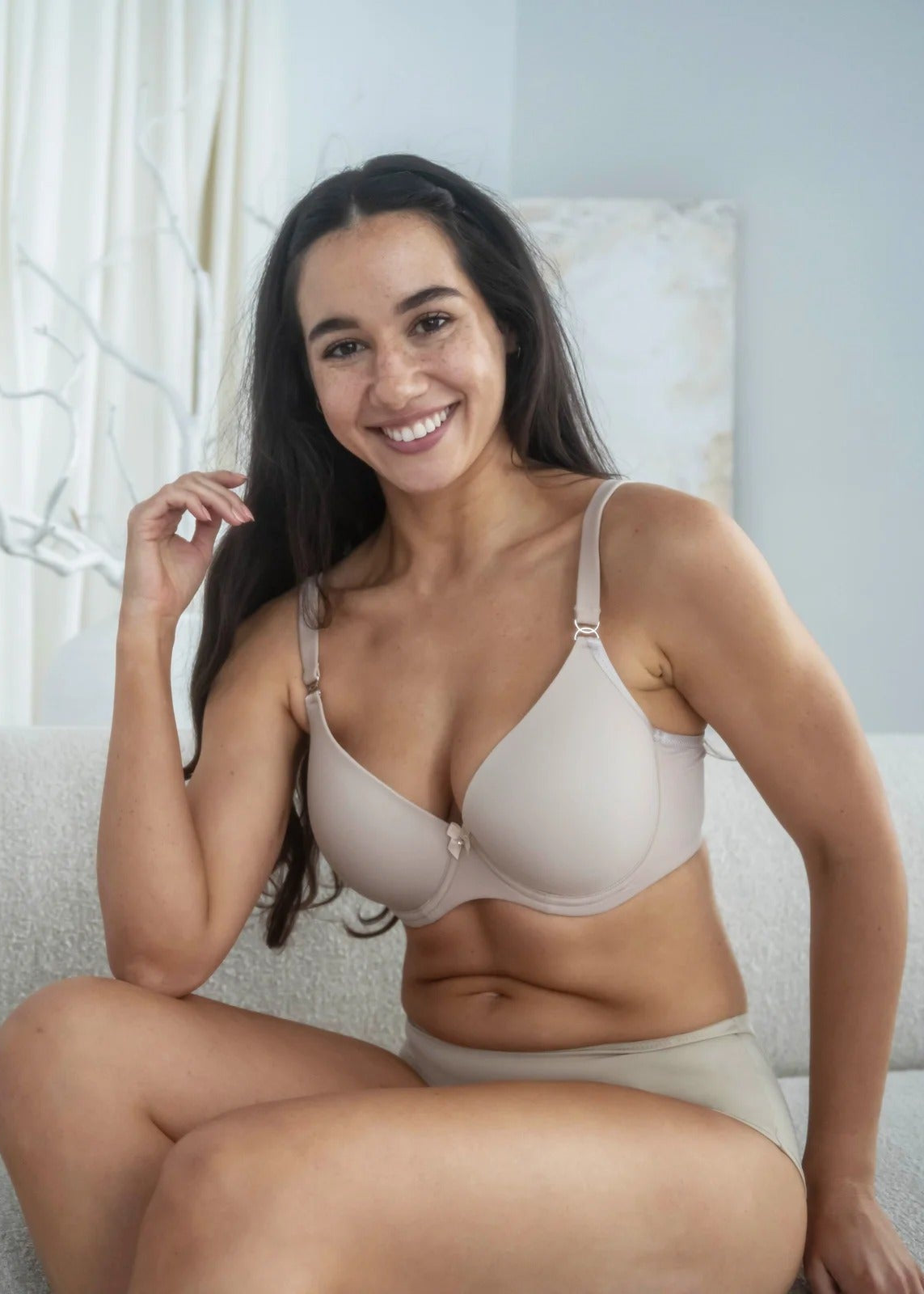 Woman wearing a beige bra and panties sitting on a couch in a bright room.