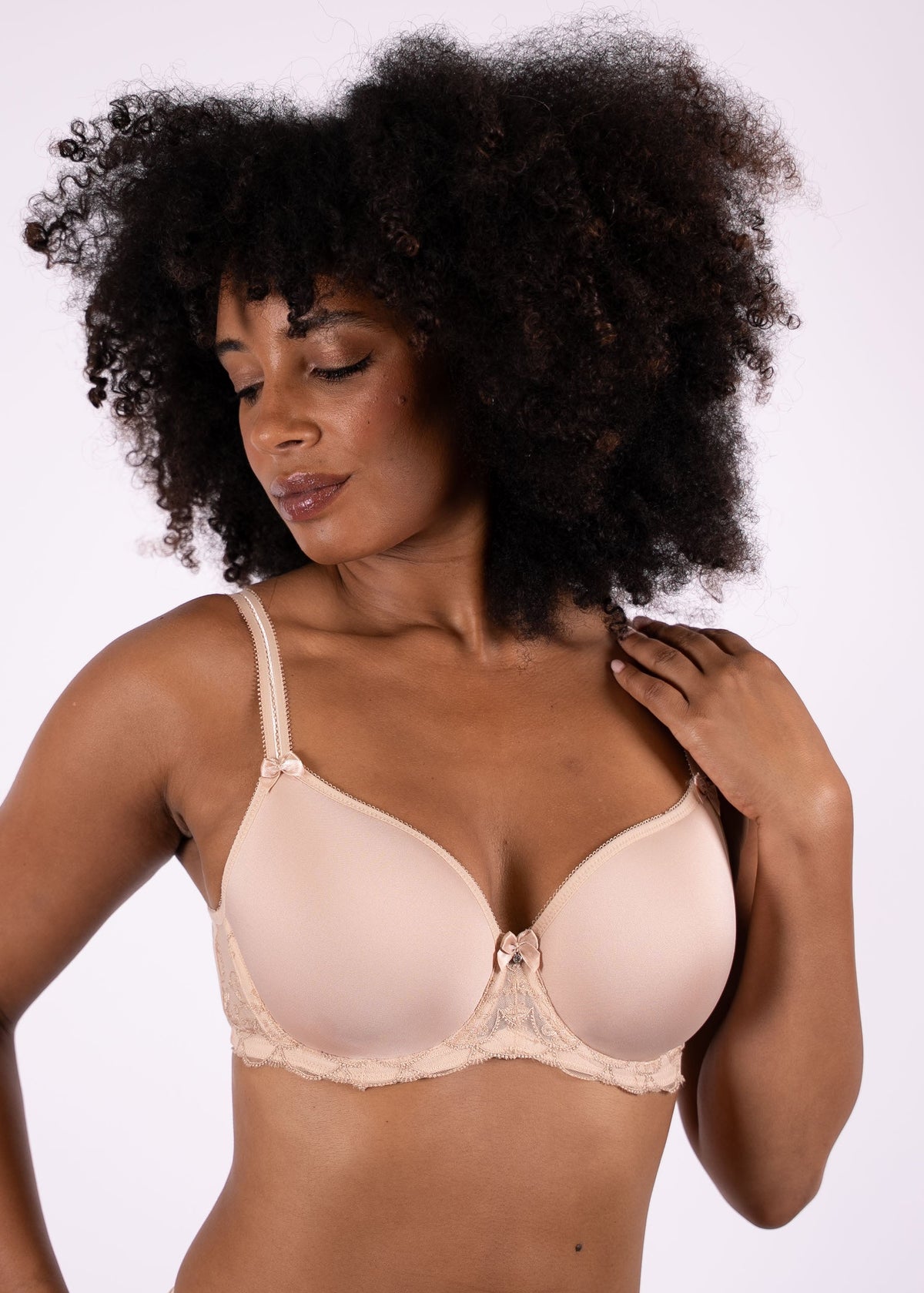 A woman with voluminous dark curly hair models a light beige underwire bra against a plain light gray studio background.