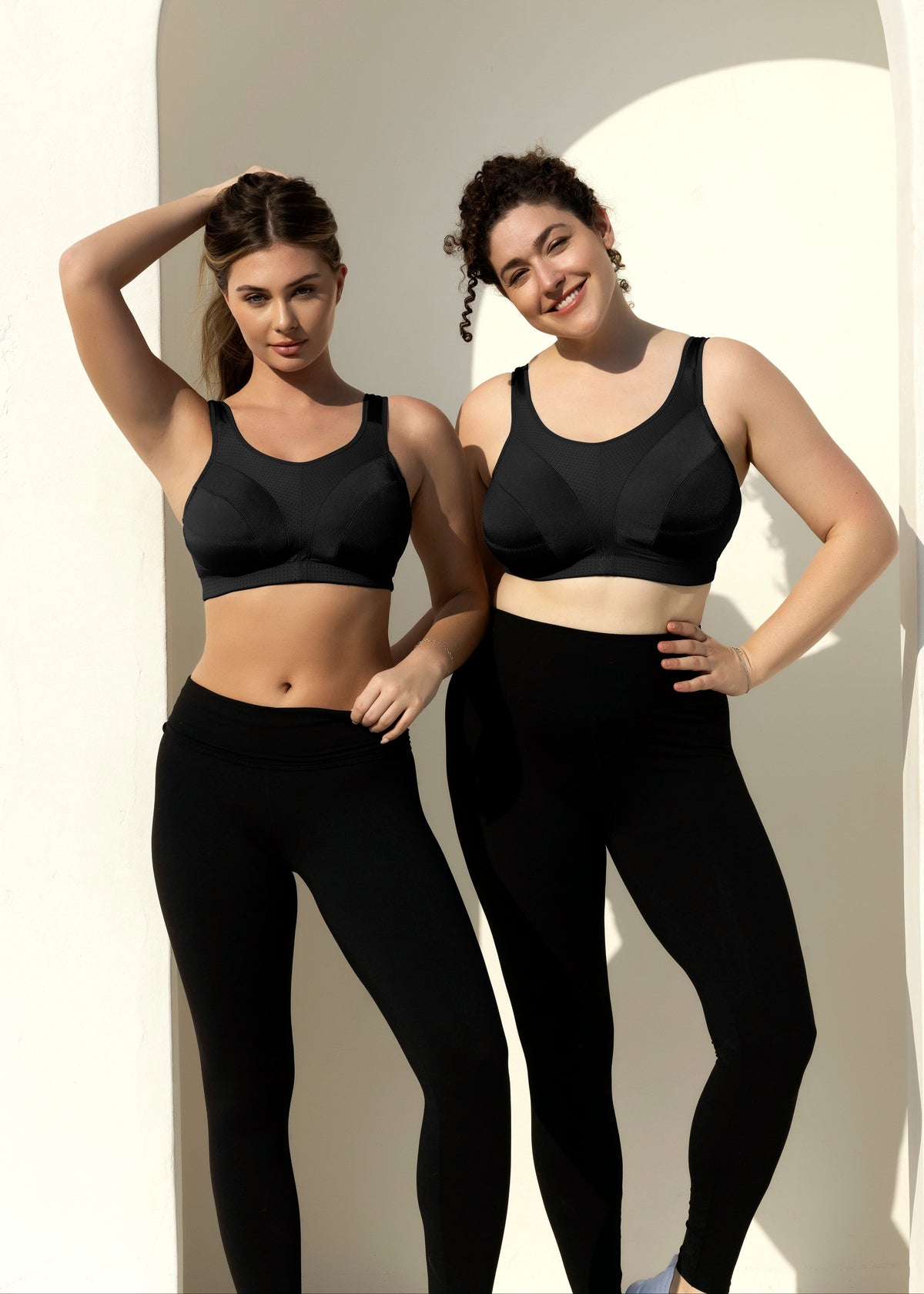 Two women modeling matching black activewear sets consisting of sports bras and high-waisted leggings, standing in front of a white arched wall.