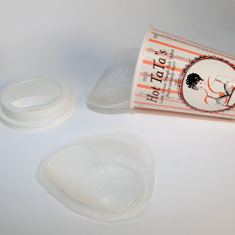 A disposable paper coffee cup with a printed illustration and the text 'Hot La Ta's' lies on its side, with a white lid and two clear plastic sleeve liners placed beside it on a white surface.