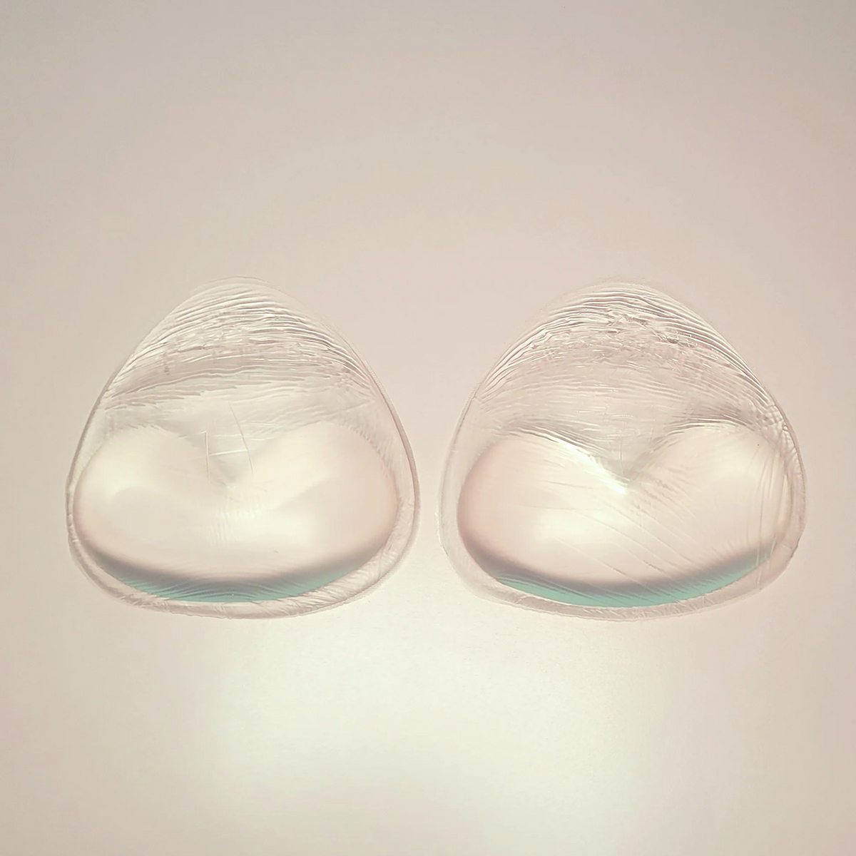 Two clear silicone bra inserts with a textured top layer and a contoured white inner cup shape, displayed side-by-side on a light beige background.