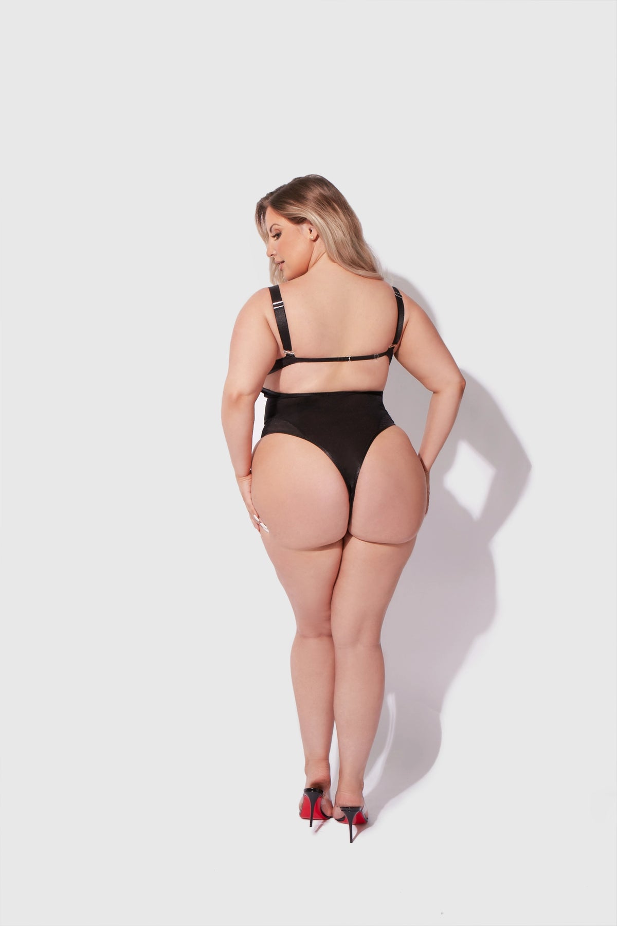 back view of a plus sized Woman wearing a black Yvone sheer shimmer mesh underwire bodysuit with demi cups, adjustable straps and high-cut tanga bottom.